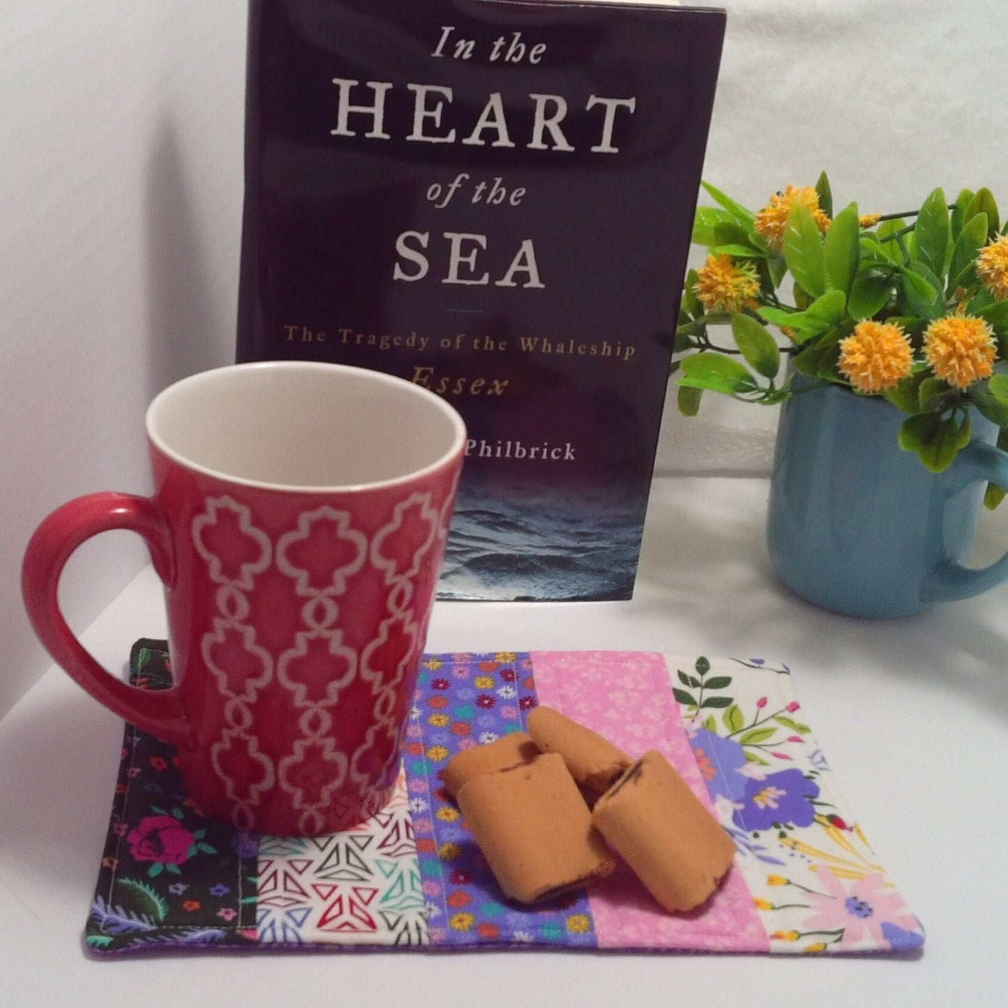 Handmade quilted mug rug in modern colors of black,purple, and bright pink.Floral patterns with mixed geometric pattern. Featured showing a book and floral arrangement in the background and a mug with cookies on the mat. Mat only for sale.